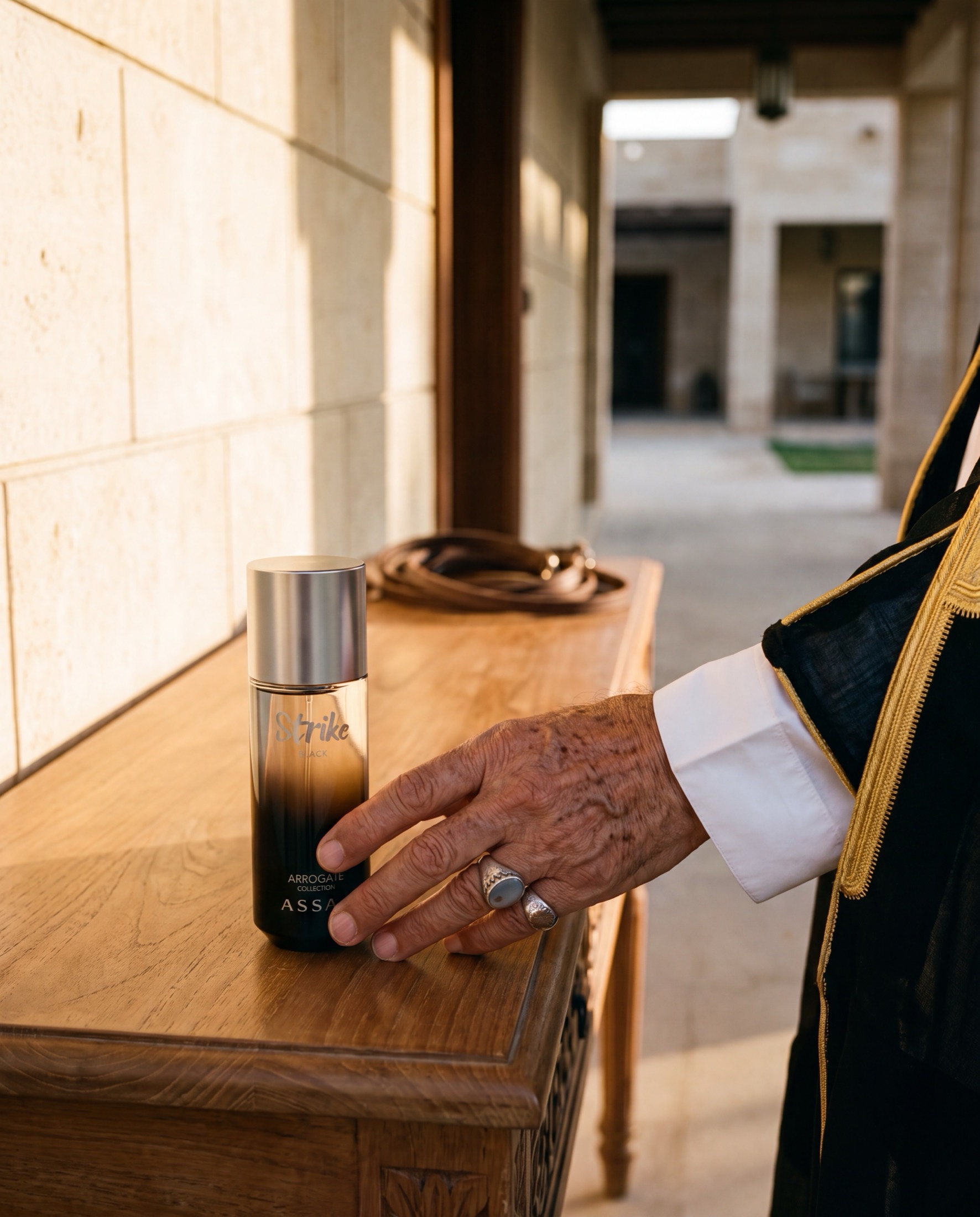 An older man's hand, wearing an agate signet ring and a dark bisht over a white cuff, resting beside an ASSAF Strike Black bottle on a carved wooden console.