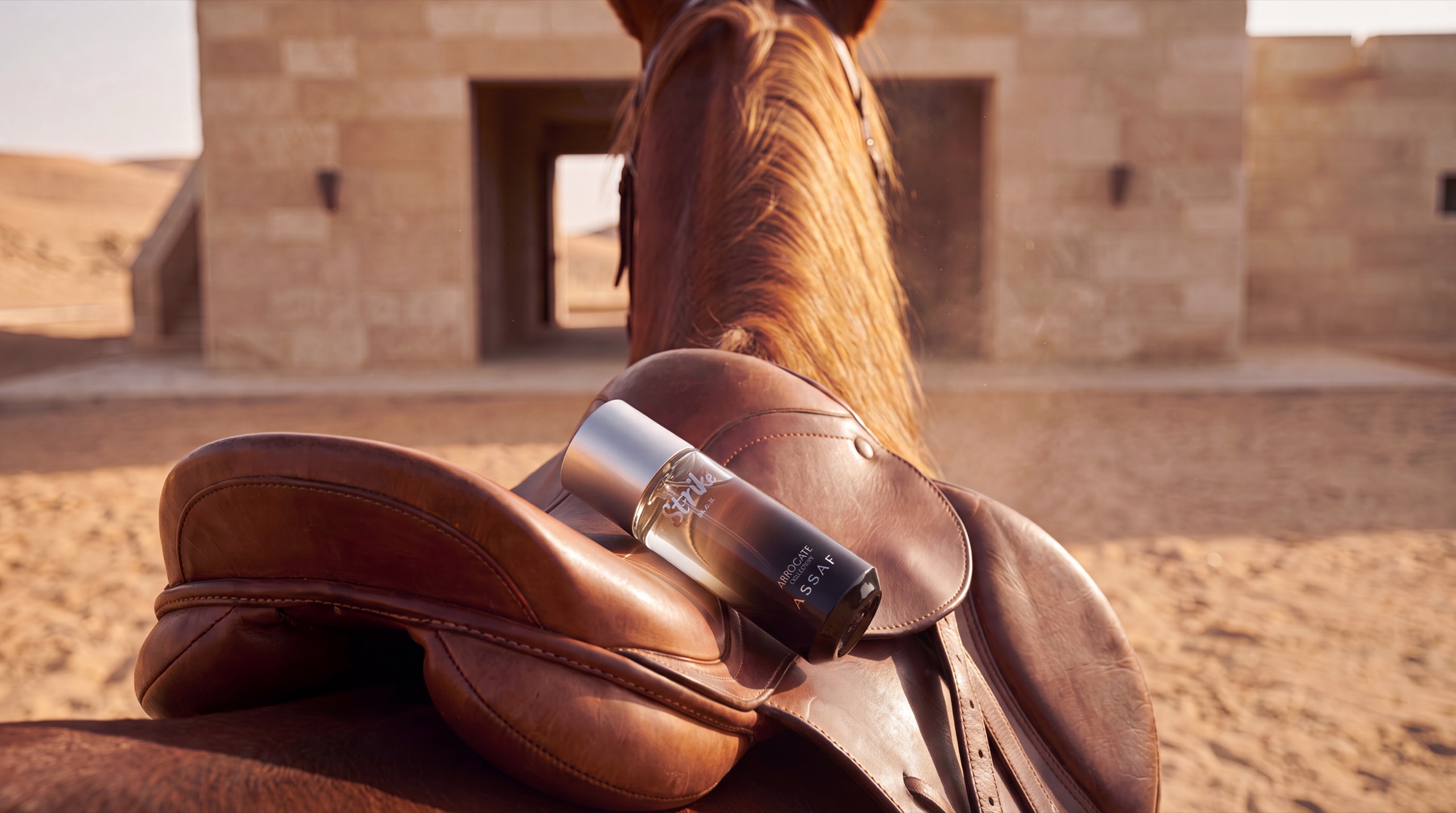 An ASSAF Strike Black bottle resting in the seat of a tan leather saddle on a chestnut horse's back, a sandstone stable and dunes behind.