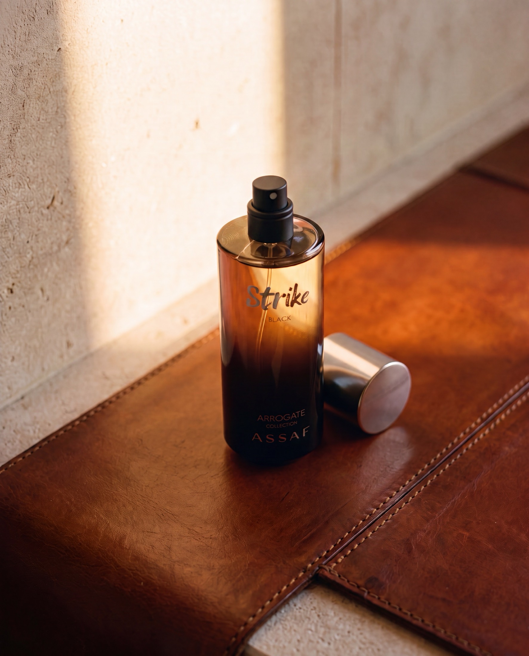 An uncapped ASSAF Strike Black bottle standing on a tan leather surface beside its brushed metal cap, warm light falling across a travertine wall.