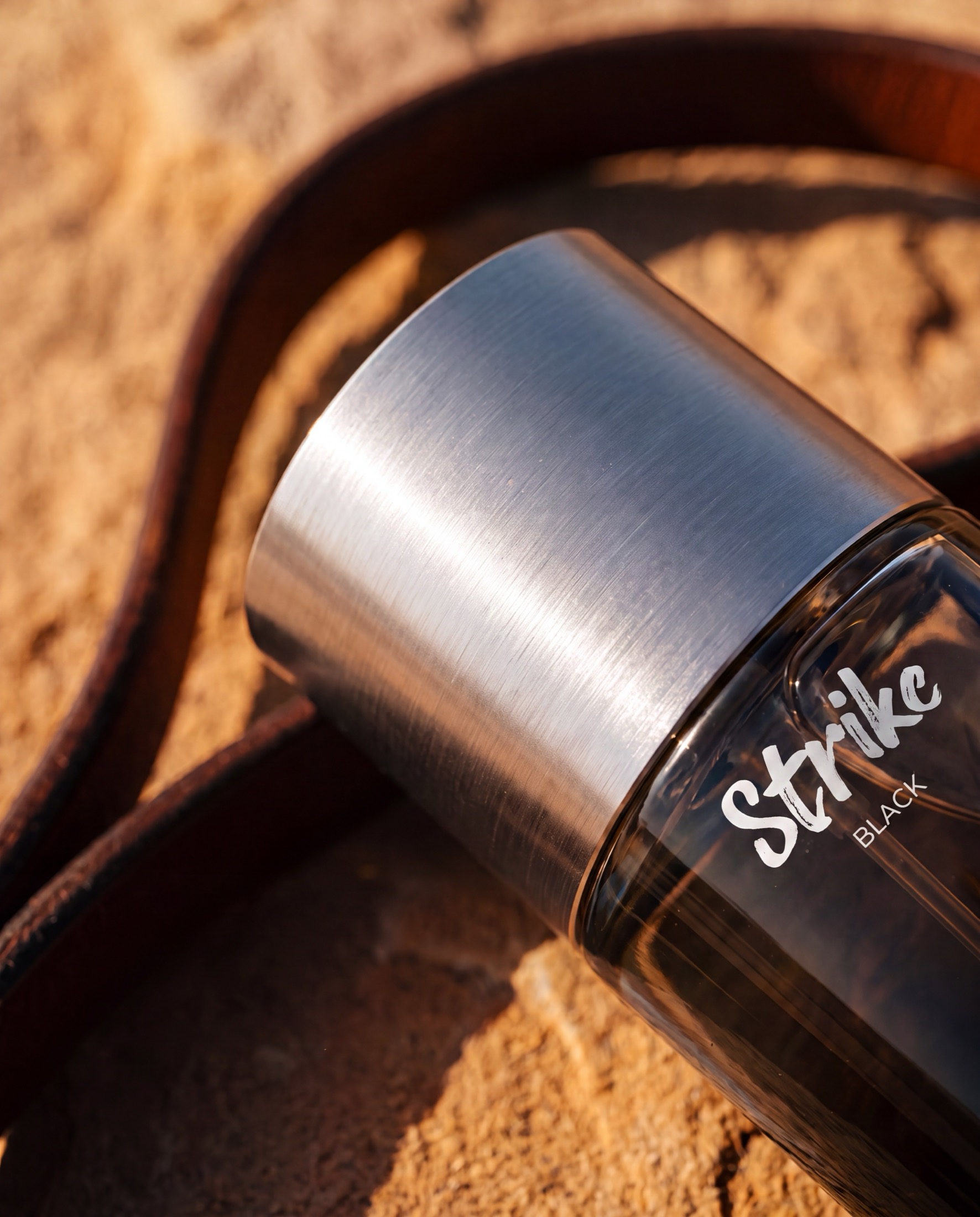 A macro view of the brushed metal collar of an ASSAF Strike Black bottle meeting the glass shoulder, a leather rein coiled on the sand behind.