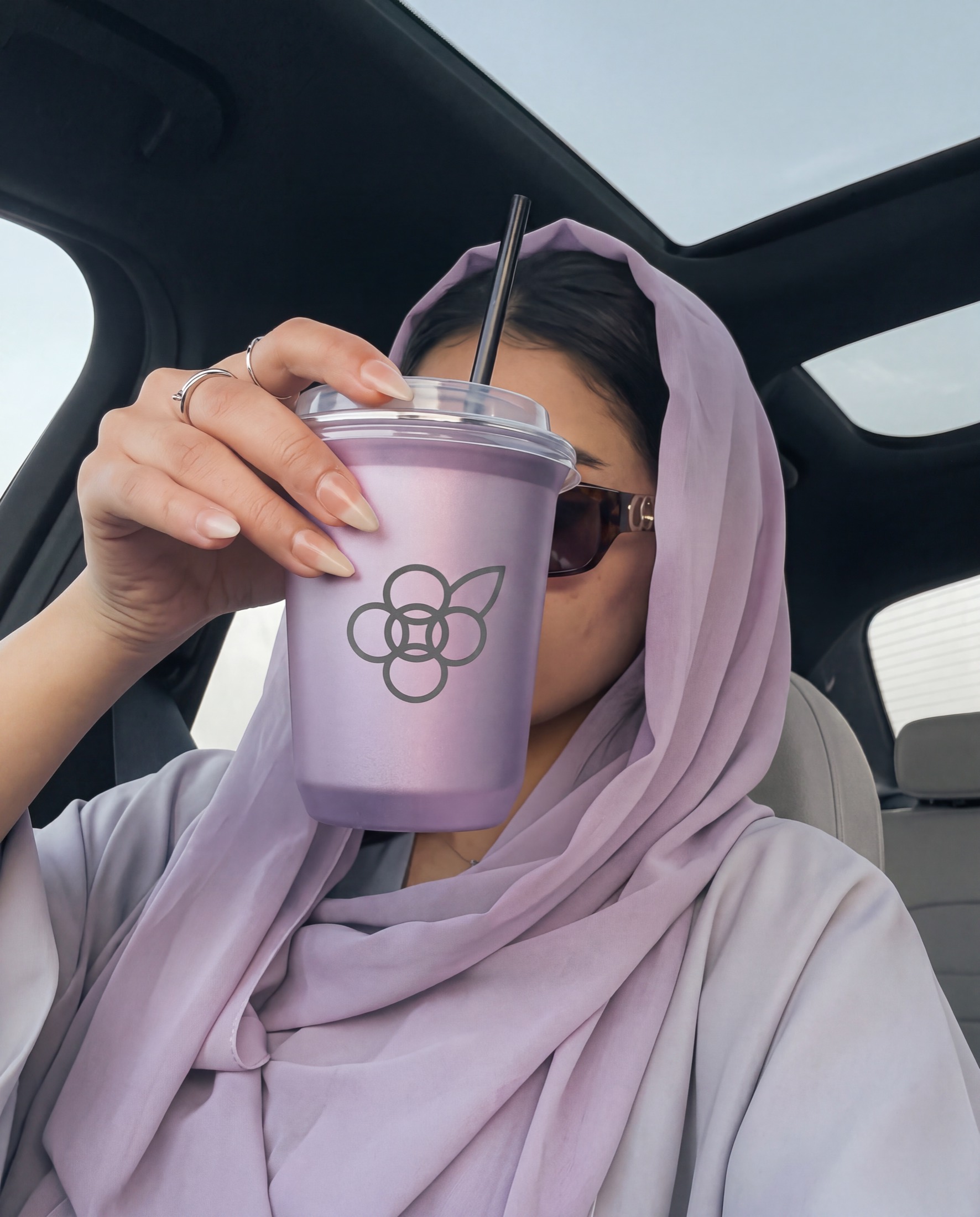 A woman in a lilac hijab sitting in a car, holding a lilac OMatcha cup with a straw up to her face.
