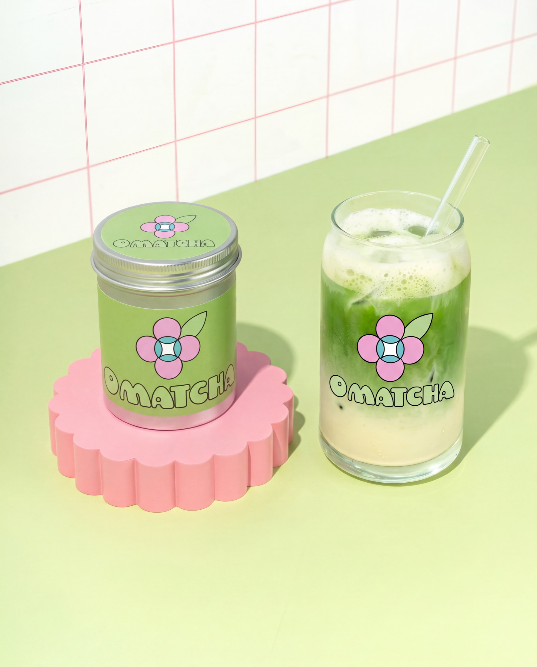An OMatcha jar on a pink flower-shaped coaster beside a glass of iced matcha, on a pale green surface with tiled wall behind.