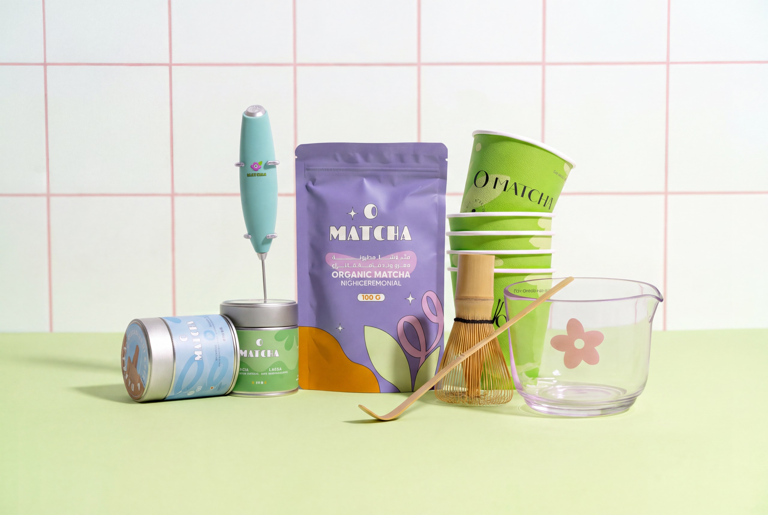 The OMatcha line arranged together on a pale green surface — two tins, a purple pouch, a milk frother, a stack of green cups, a bamboo whisk and scoop, and a glass jug.