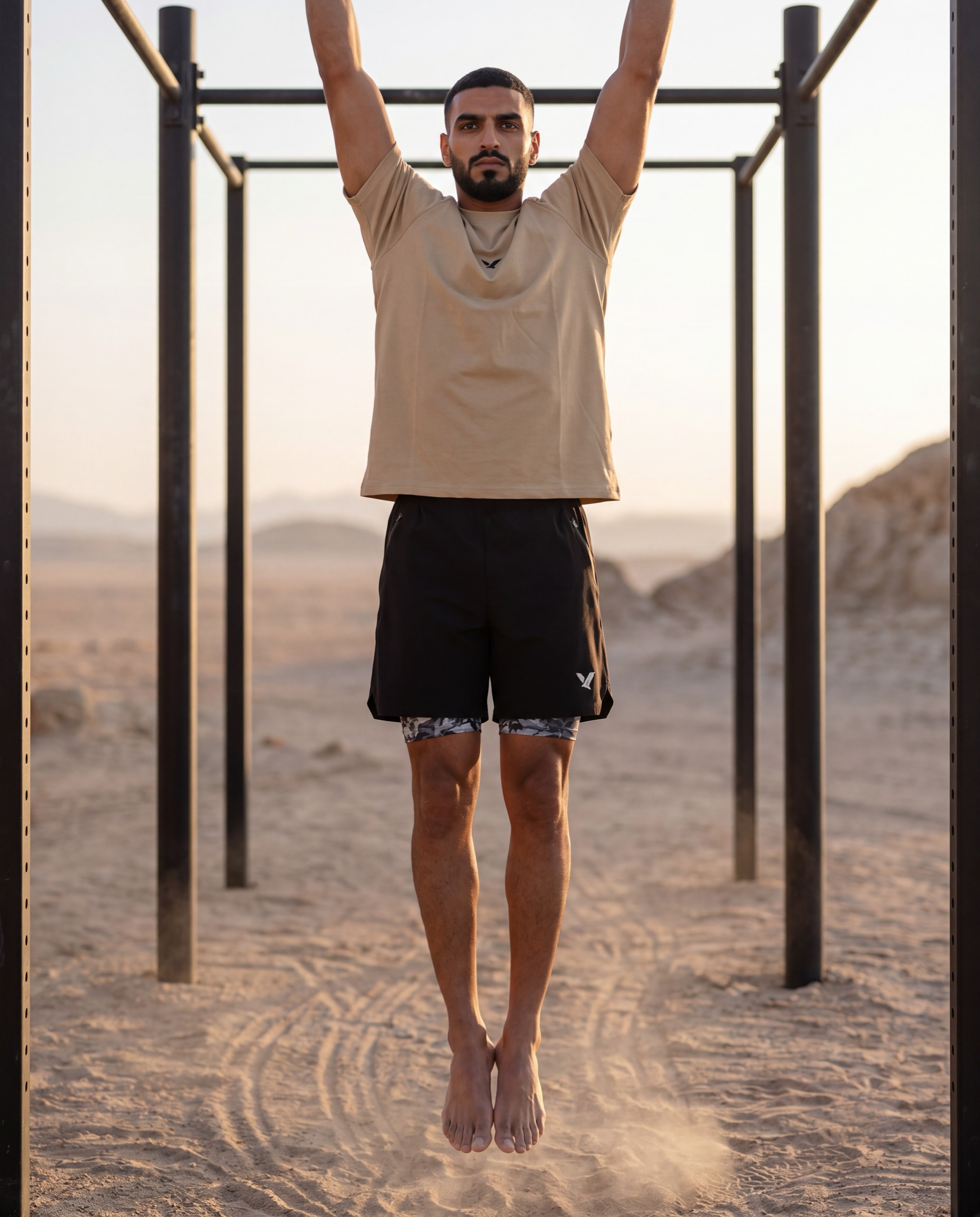 A man in an OQAB oversized shirt hanging at full stretch from the pull-up rig, legs straight, sand beneath.