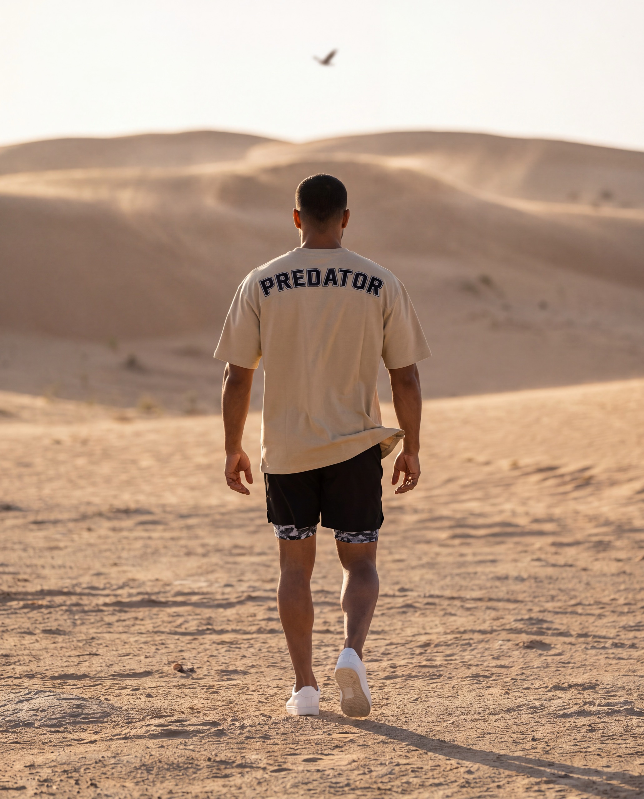 A man walking away across the sand toward the dunes, the arched PREDATOR lettering across the back of his OQAB oversized shirt, an eagle small in the sky.